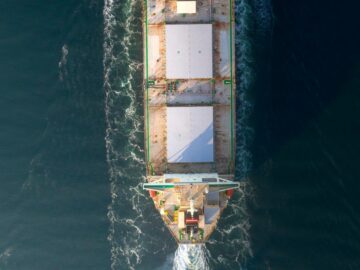 An aerial photo of a large cargo ship with tugboats assisting in open sea, showing maritime transportation.